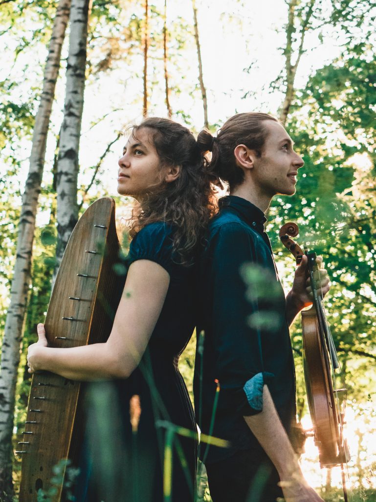 Eva Väljaots & Robbie Sherratt holding a kannel and fiddle standing back to back in a forest.